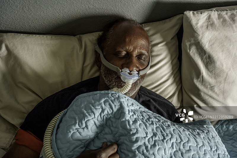 一位七十多岁的非裔美国老人安静地躺在床上，使用CPAP（持续正压呼吸）机来缓解睡眠呼吸暂停。图片素材