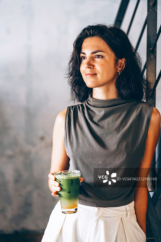 拉丁裔女性手持一杯冰抹茶。她穿着绿色无袖上衣和白色裤子。城市环境中，柔和的灯光和现代楼梯。图片素材