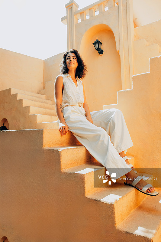拉丁裔女性穿着无袖白色服装，坐在米色楼梯上。她微微仰头，面带微笑。地中海风格的建筑在温暖的阳光下。图片素材