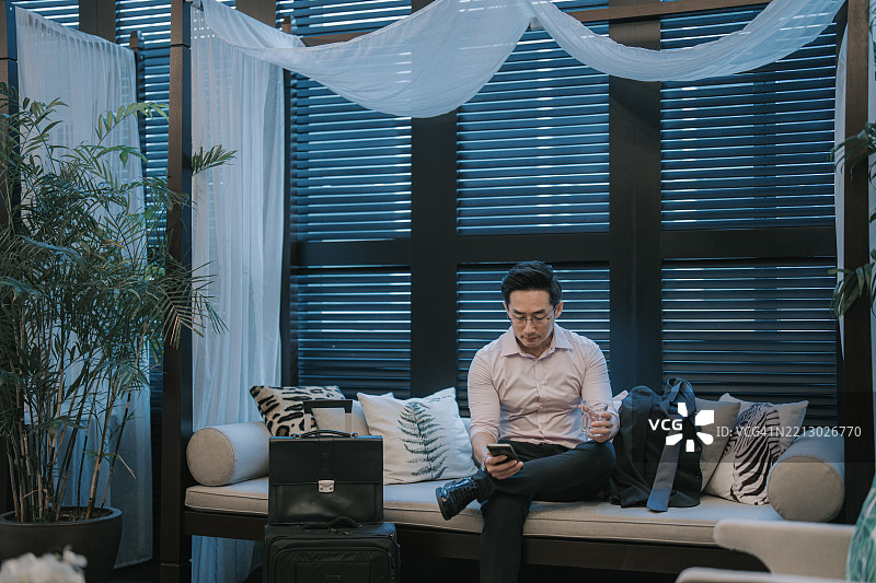 亚洲商务旅客在酒店休息室放松，手中拿着饮料和智能手机图片素材