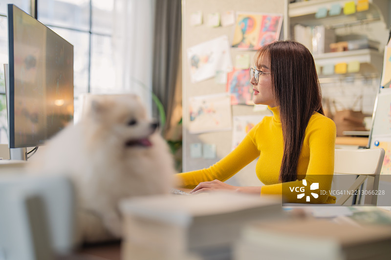 亚洲创意机构的女性穿着休闲服装，在家中工作，设计广告媒体出版，同时听着音乐，身边有她的小白色博美犬，工作室充满创意氛围。图片素材