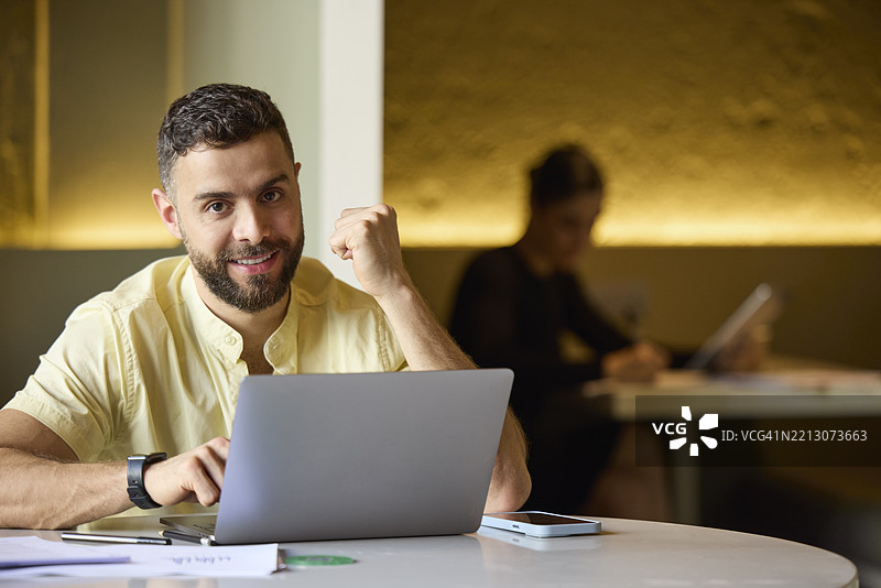 微笑的男性自由职业者坐在办公室里，握拳庆祝成功。快乐的商人在庆祝胜利，桌上有一台笔记本电脑。图片素材
