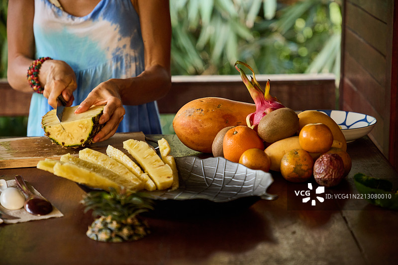一个人在浅色木桌上切菠萝，周围摆放着新鲜的热带水果图片素材