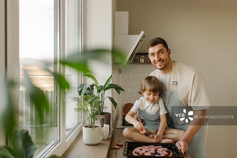 父亲和儿子在明亮的厨房里一起烹饪，阳光透过窗户洒入。图片素材