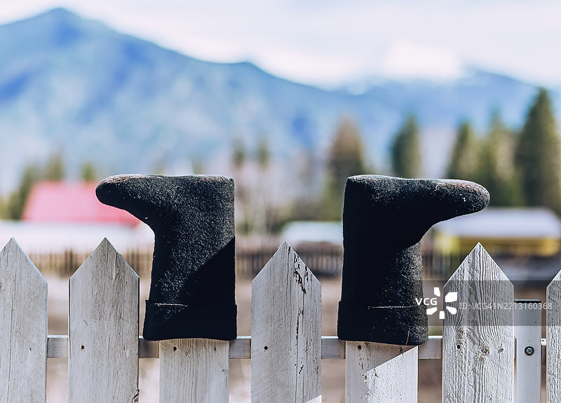 白色围栏上的黑色毛毡靴，背景是山脉。成双的黑色毛毡靴在白色木栅栏上晾干，背景是美丽的山脉和树木。夏季圣诞，幽默。图片素材