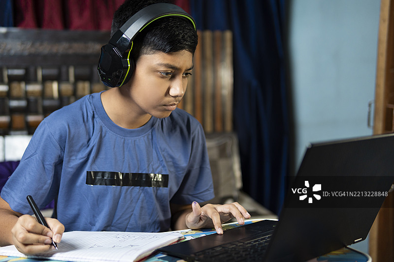 印度男孩戴着耳机在家用笔记本电脑在线学习，写笔记，做作业。图片素材
