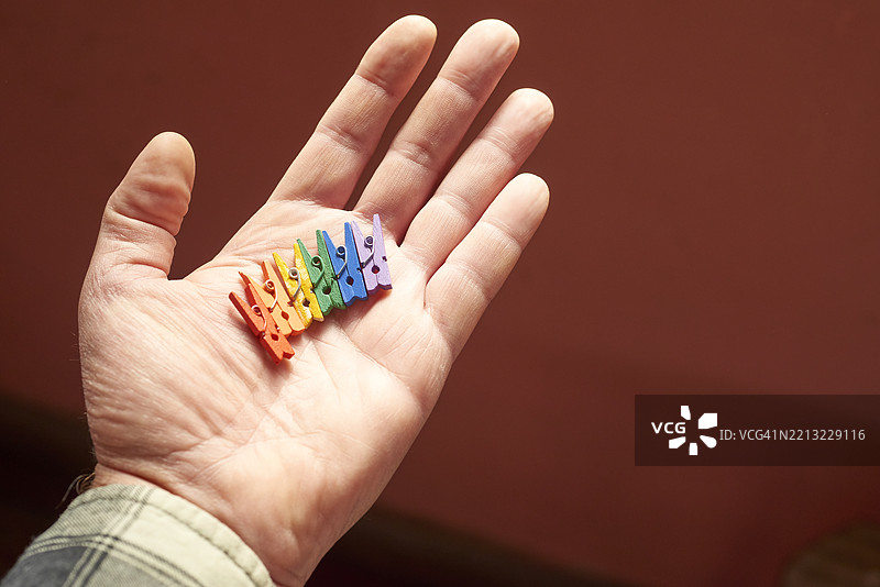白人手将五颜六色的衣夹以彩虹形状展开, resting against warm brown surface,象征着创意排列和色彩多样性。图片素材