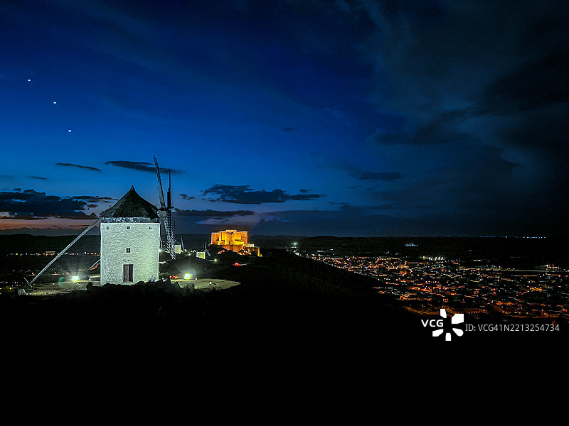 在西班牙卡斯蒂利亚-拉曼查的孔苏埃格拉,传统的风车(堂吉诃德的风车)在日落后黄昏时分排列整齐并被照亮,背景是孔苏埃格拉城堡和戏剧性的天空。图片素材
