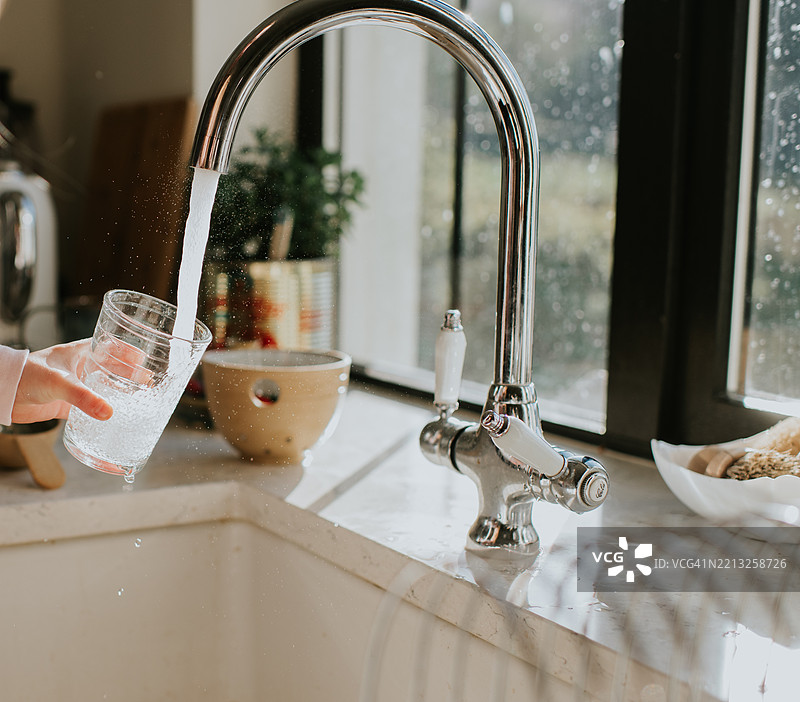 阳光下，年轻孩子手持玻璃杯，水从闪亮的水龙头流入杯中，旁边是现代厨房水槽。图片素材