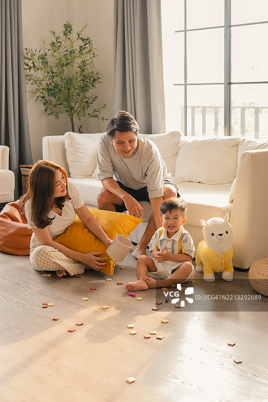 年轻亚洲家庭的肖像，父亲、母亲和幼儿。亚洲家庭坐在客厅里，父母和孩子在家中度假时放松。快乐的母亲在家中的木地板上与可爱的小男孩玩耍。图片素材