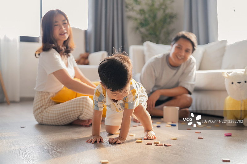 年轻亚洲家庭的肖像，父母和幼儿坐在客厅里，父母和孩子在家中度假时放松。快乐的母亲在家中的木地板上与可爱的小男孩玩耍。图片素材
