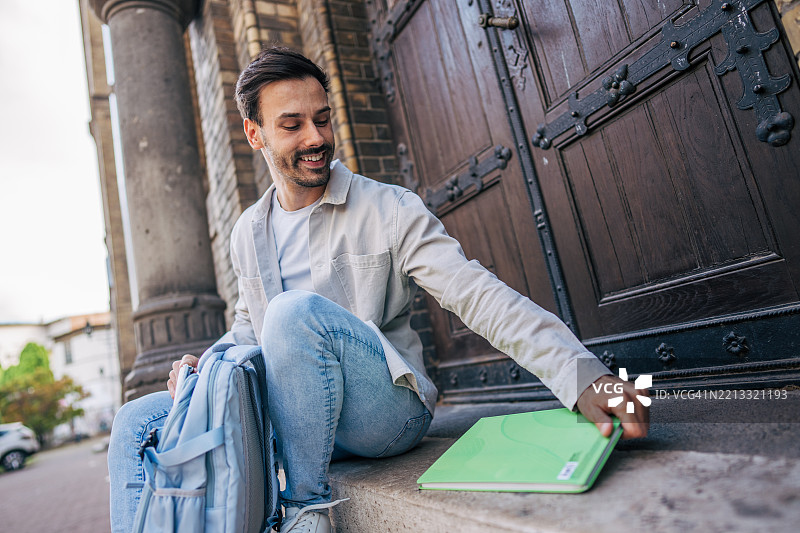 微笑的学生在大学建筑外捡起一本书图片素材