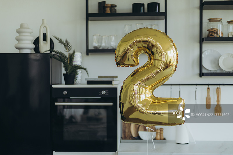 光滑的金色铝箔气球，形状为数字2，漂浮在厨房里。图片素材