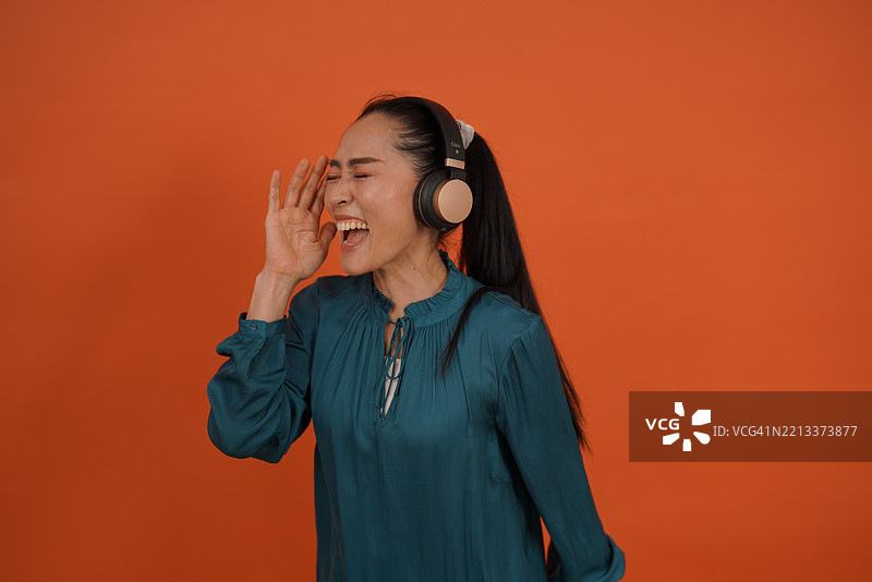 亚洲女演员穿着橙色长袖衫，戴着耳机和使用手机的有趣照片。图片素材