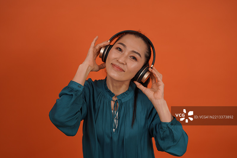 亚洲女演员穿着橙色长袖衫，戴着耳机和使用手机的有趣照片。图片素材