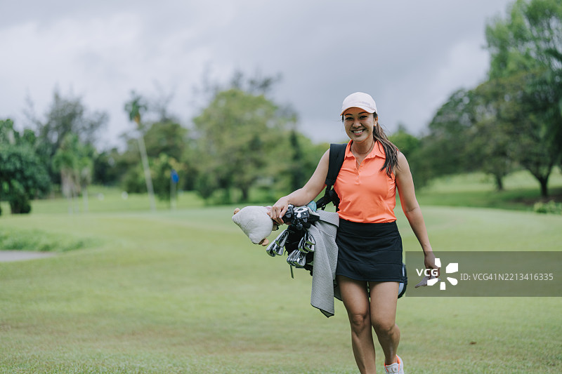 微笑的亚洲女性高尔夫球手背着高尔夫球包走过球道图片素材