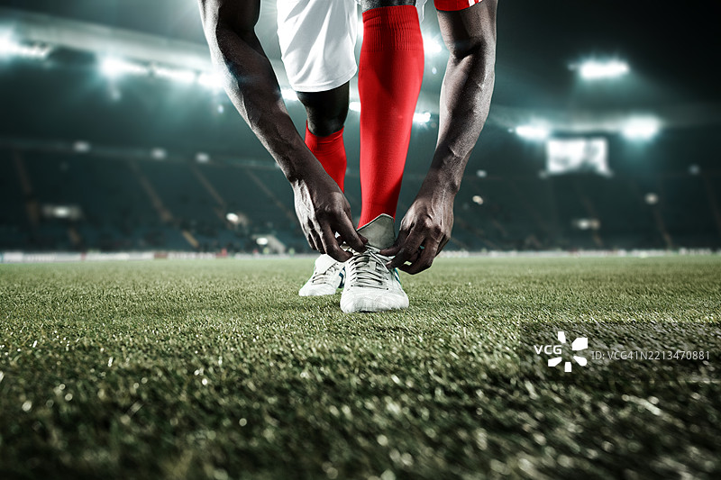 合成图像。裁剪的照片显示一名穿着白色短裤和红色袜子的男性足球运动员在绿色草地上站着，正在系足球鞋的鞋带。图片素材