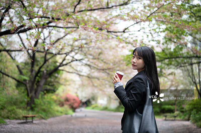 一位年轻的日本女性手握咖啡杯，在樱花盛开的公园中回头望去。图片素材