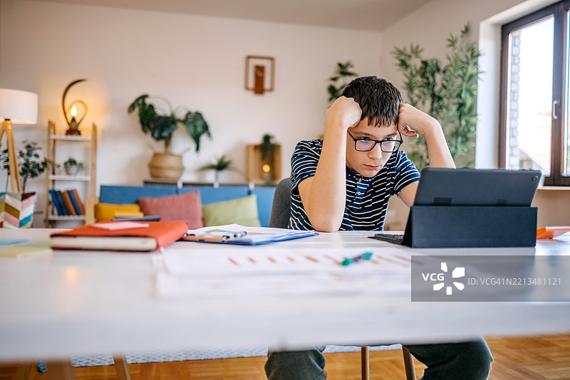 年轻的少年在家中使用平板电脑学习，坐在餐桌旁。图片素材