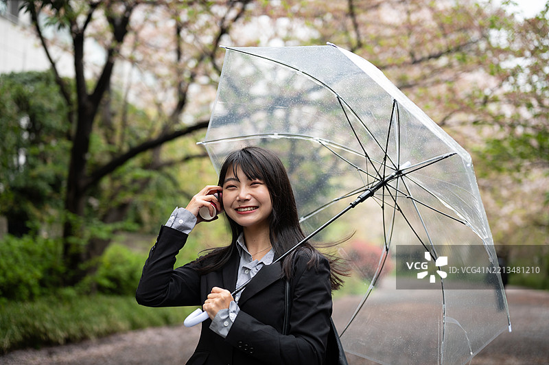 一位日本女性微笑着，手里拿着一个空咖啡杯，站在樱花树下的公园里，撑着一个塑料伞。图片素材