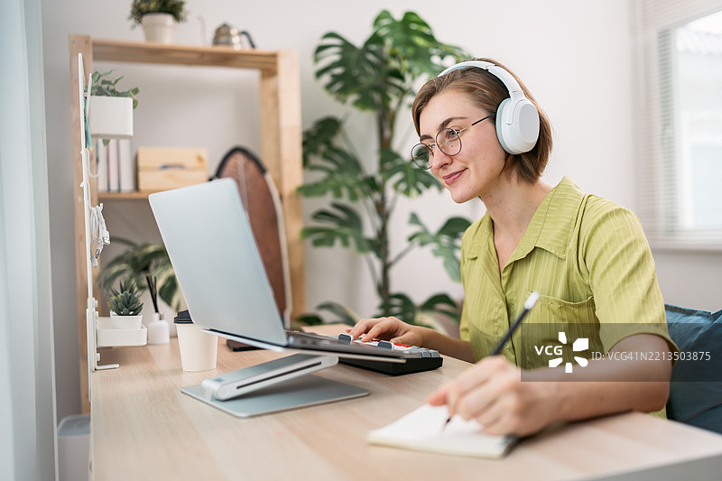 快乐的年轻女性在家中一边戴着耳机，一边使用笔记本电脑和记事本进行多任务处理图片素材
