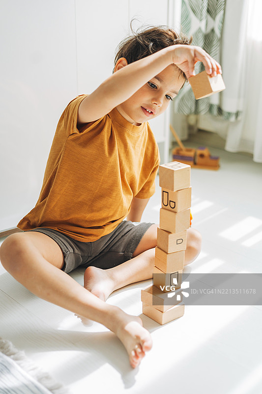 一个专注的小男孩在地板上小心地堆叠木块，正在建造一座塔。图片素材