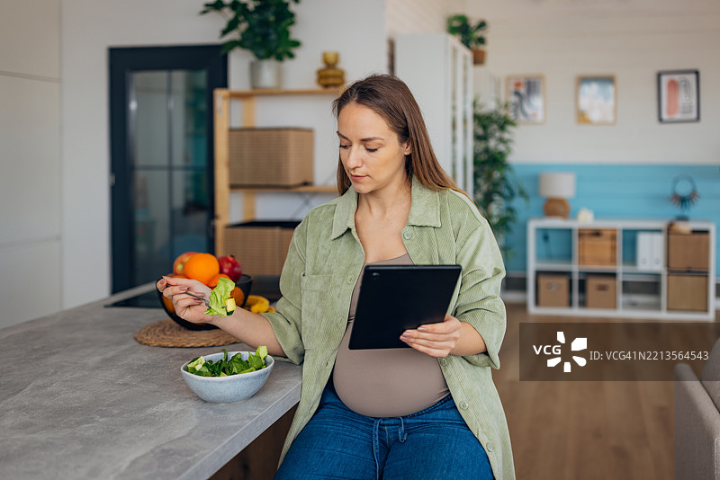 微笑的孕妇在厨房吃早餐时使用平板电脑图片素材