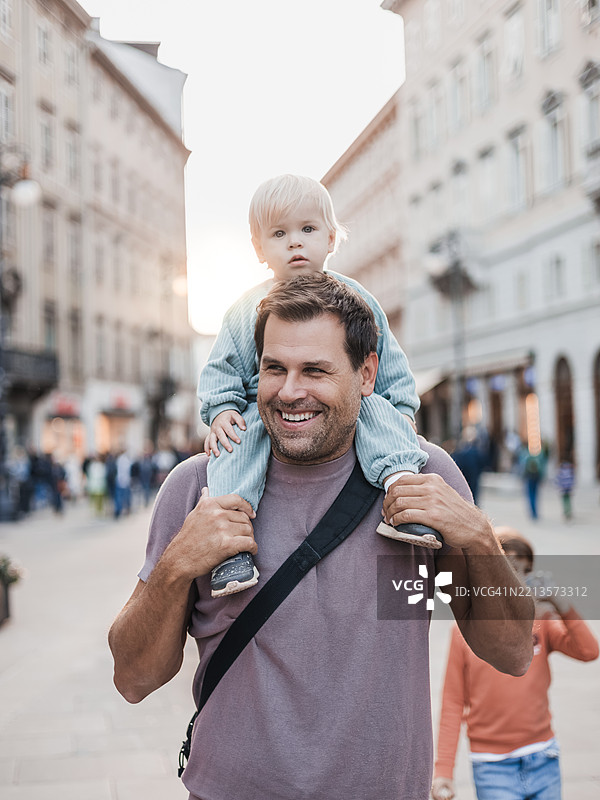 微笑的父亲在城市中背着他的幼儿，享受家庭假期。图片素材