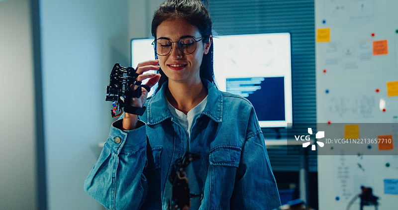 年轻女性工程师在夜间的科技实验室加班，手势指向机械手臂。机器学习、计算机制造、创新与创造力。人工智能与机器人开发。图片素材