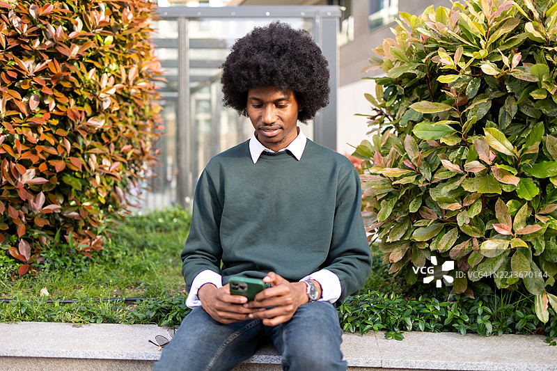 男人坐在户外，使用智能手机，周围环绕着植物和绿意。图片素材