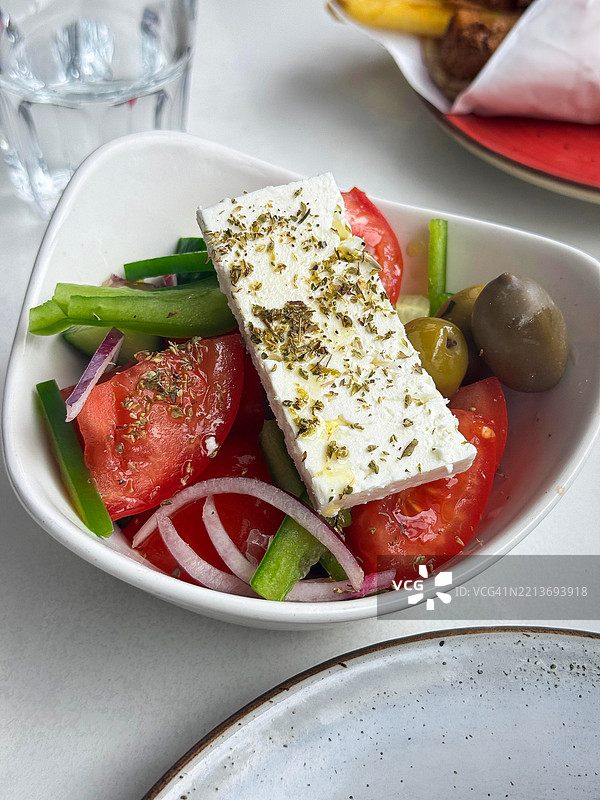 白色碗中盛着希腊沙拉（Horiatiki），配有浸泡在橄榄油和干牛至中的菲达奶酪块，沙拉包含切片番茄、青椒、红洋葱、绿橄榄和切碎的黄瓜，背景为白色桌布，俯视角度。图片素材
