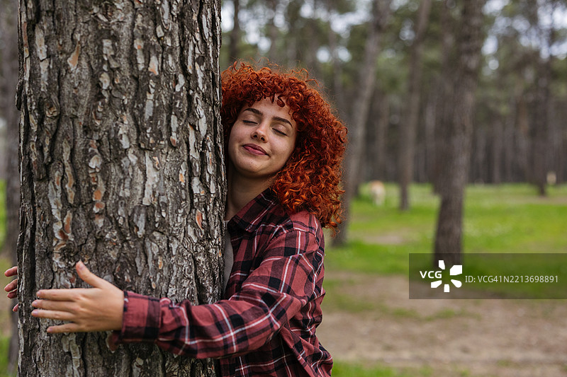 年轻女子在森林中拥抱一棵树，红色头发。图片素材