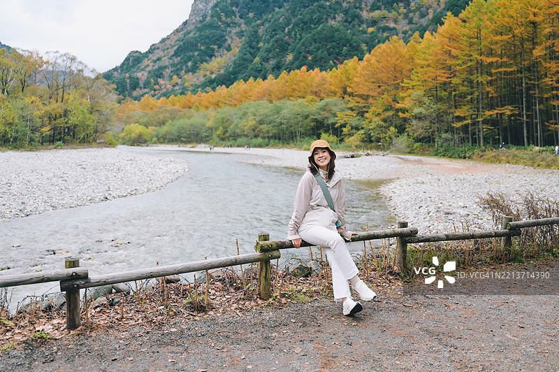 在上高地河边小径旁摆姿势的年轻女性，享受放松的旅行时刻，生动的秋季色彩和山脉风景，日本阿尔卑斯的自然旅游。图片素材