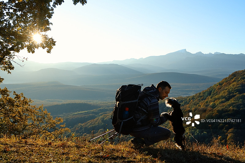 探险者在山间徒步旅行时向狗打招呼。阳光照耀的秋季山谷在这对徒步者的脚下延展。图片素材