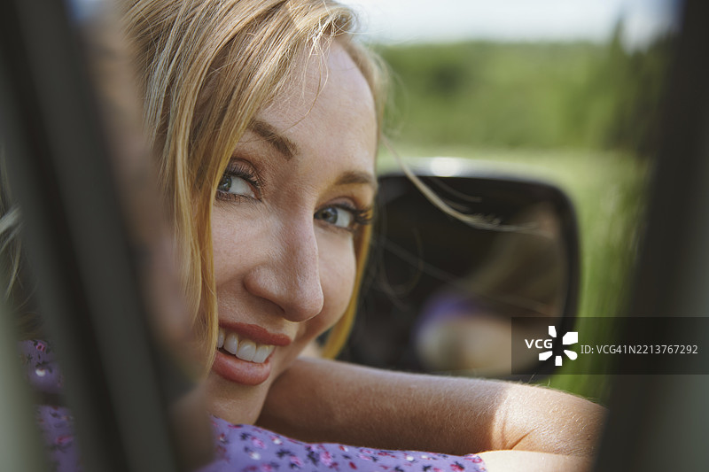 金发女子在夏季公路旅行中，从车窗微笑，背景是一片绿色的田野，美国，北美。图片素材
