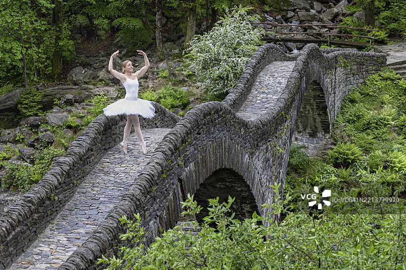 穿着白色芭蕾舞裙的芭蕾舞者在弯曲的石桥上优雅地摆姿势,周围是绿色的自然环境,地点为瑞士提契诺州洛卡尔诺区的维尔扎斯卡谷的萨尔蒂桥。图片素材