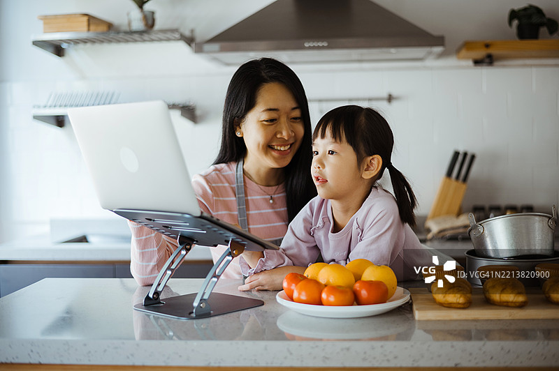 亚洲母亲在厨房里用笔记本电脑帮助她的小女儿学习图片素材