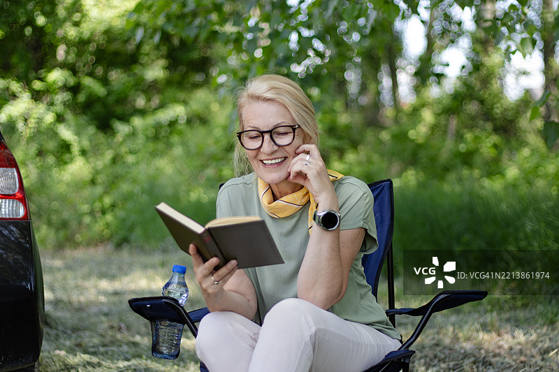 一位成熟的金发女性在度假时来到大自然，她享受坐在椅子上阅读一本书的时光。图片素材
