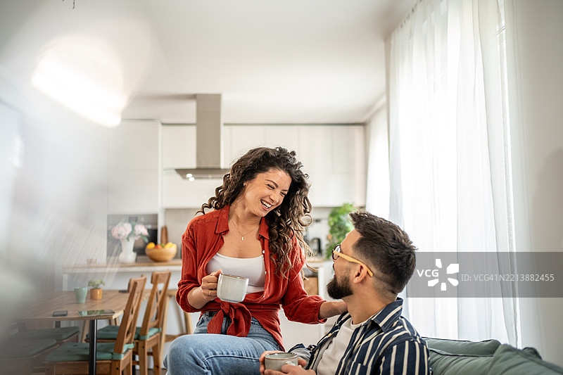 年轻情侣在沙发上放松，喝着咖啡，开心地笑着图片素材
