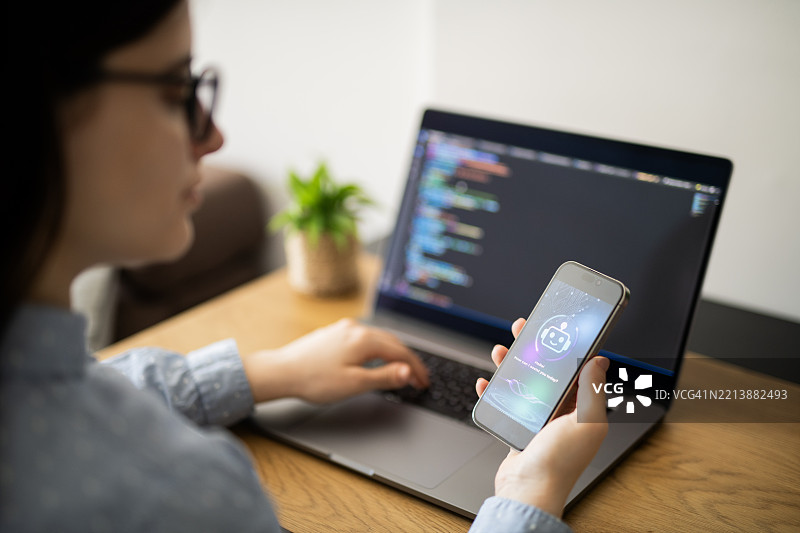 IT程序员正在使用智能手机上的AI聊天机器人应用程序来辅助她在笔记本电脑上工作。图片素材