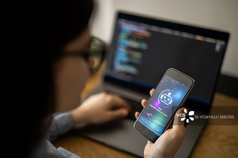 IT程序员正在使用智能手机上的AI聊天机器人应用程序来辅助她在笔记本电脑上工作。图片素材