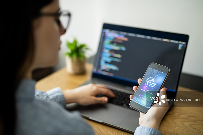 女商人在使用智能手机上的AI聊天机器人应用程序，以帮助她在笔记本电脑上工作。图片素材