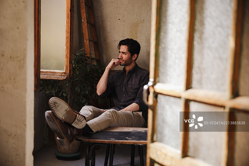 一位留着三天胡须的帅气男士坐在窗边望着外面。图片素材