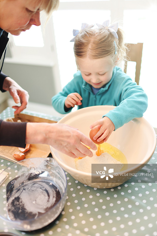 四岁的小女孩在奶奶的帮助下学习如何将鸡蛋打入一个大搅拌碗中，他们一起准备海绵蛋糕的混合物。图片素材