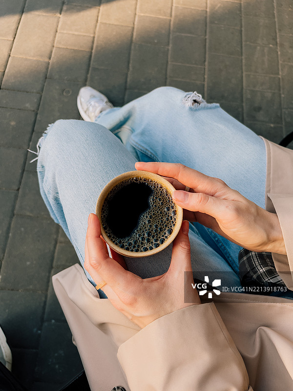 第一人称视角的女性穿着牛仔裤和米色风衣，手里拿着一杯热黑咖啡。图片素材