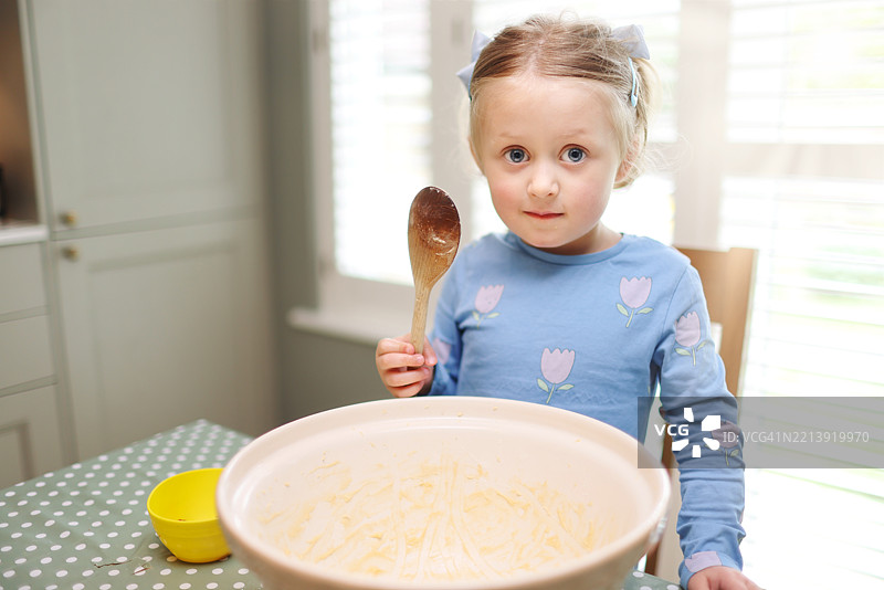 四岁女孩用木勺刮取大陶瓷搅拌碗中海绵蛋糕混合物的最后残余并品尝。图片素材