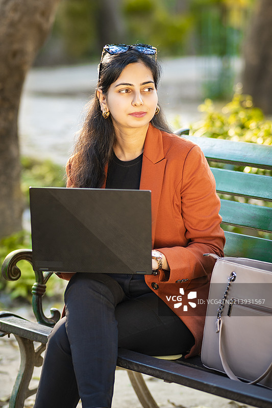 在公园长椅上微笑的印度年轻女商人户外肖像，边思考边使用笔记本电脑工作。图片素材
