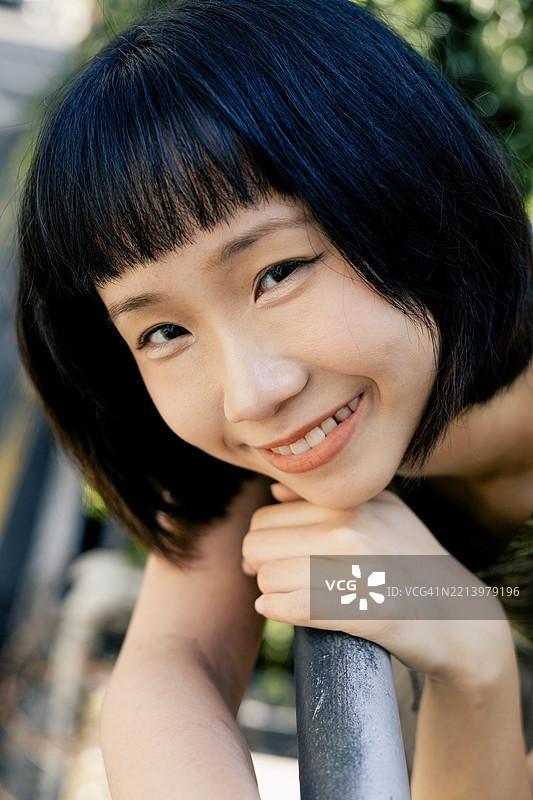 微笑的年轻亚洲女性靠在栏杆上，在自然光下看着镜头微笑图片素材