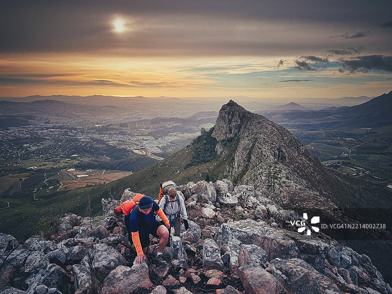 年轻充满活力的男性徒步旅行者在日落时分攀登崎岖的小径，远处是山谷和山脉的美景图片素材