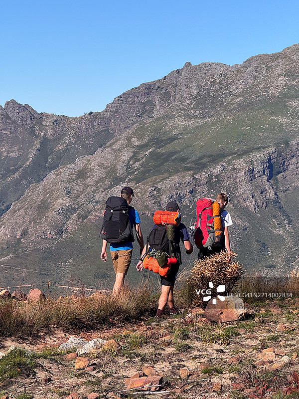 三名青少年男性徒步旅行者在晴朗的蓝天下穿越山脉附近的崎岖小径图片素材